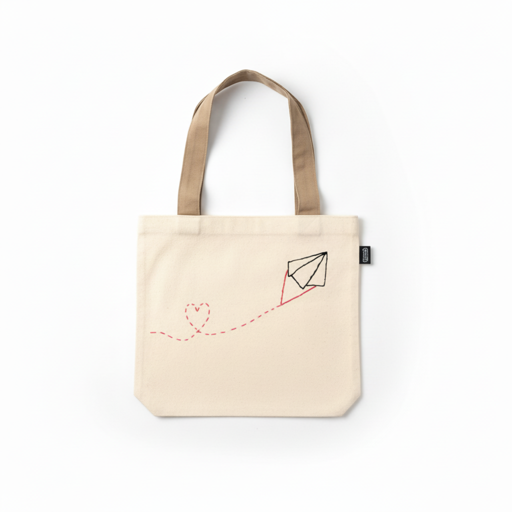 Cream beige canvas tote bag with embroidered paper airplane and heart trail on white background